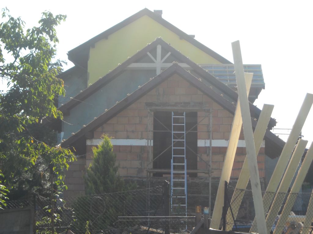 Wznoszenie murowanej ściany szczytowej z otworem okiennym, widoczna drabina rusztowania i drewniane podpory, częściowo otynkowana ściana budynku w tle.